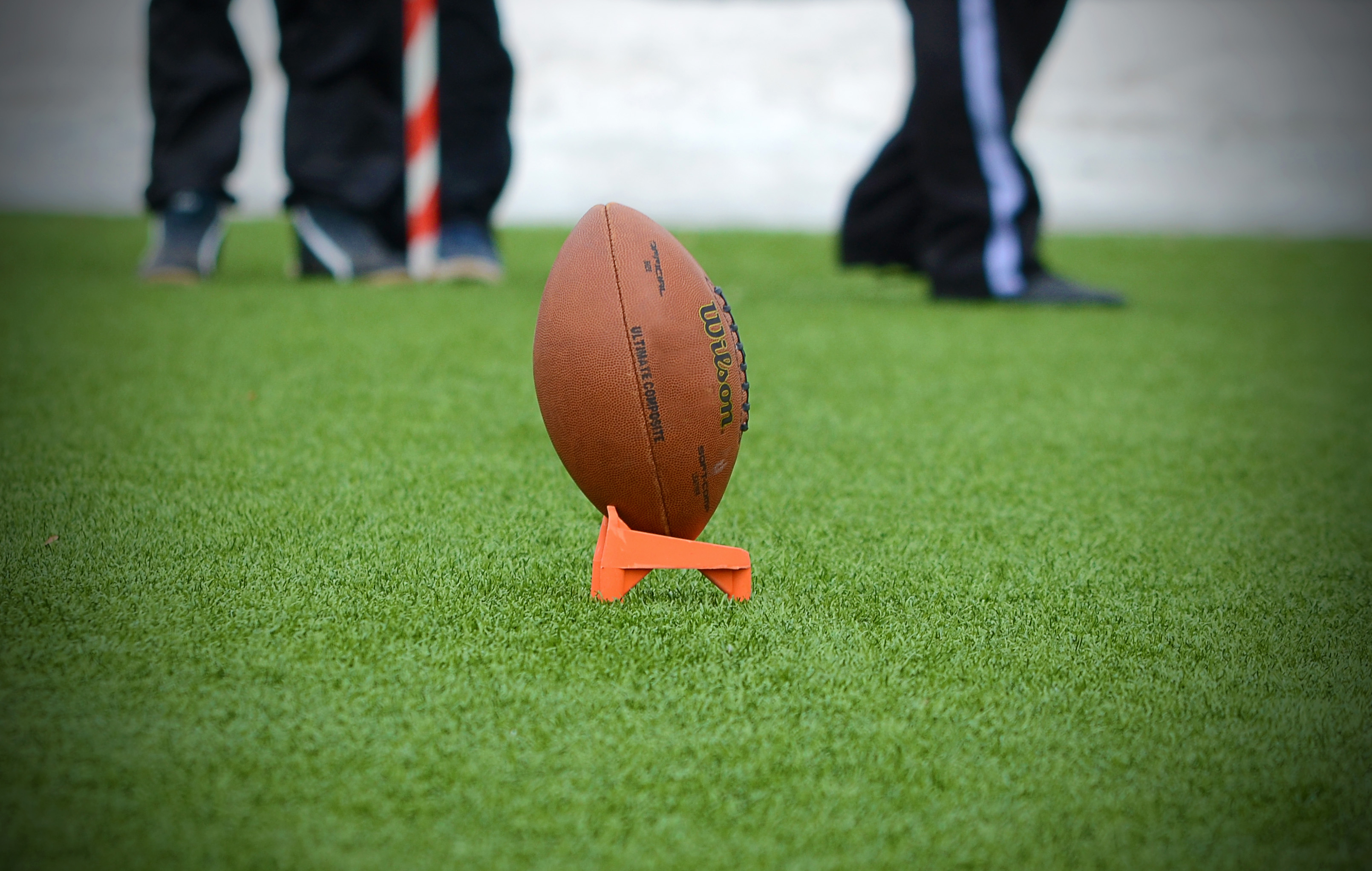 Picture of a football on a grassy field with people's legs visible in the background